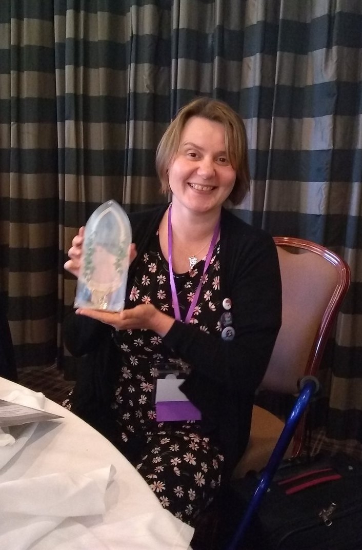 Ruth EJ Booth sat at a round dinner table in front of a gingham curtain holding the British Fantasy Award for Best Non-Fiction