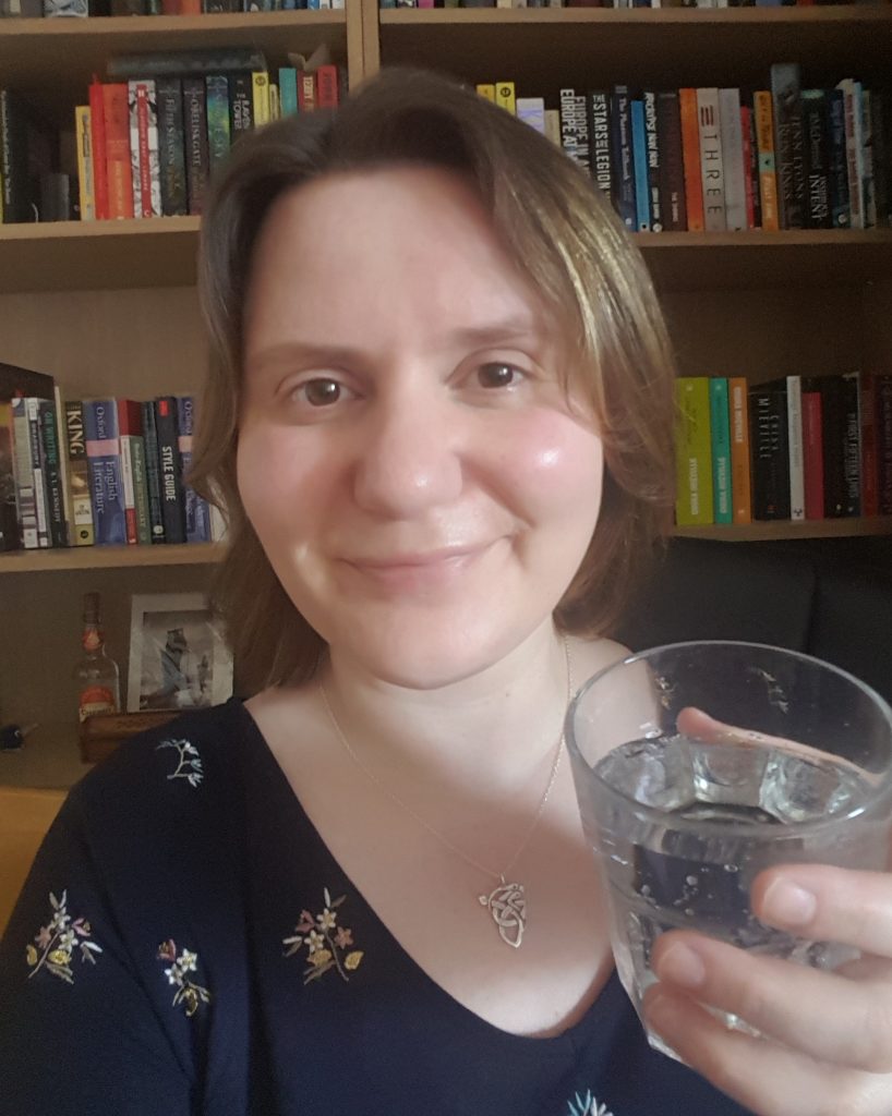 Ruth Booth holding a glass of lemonade, smiling, in front of some bookshelves.