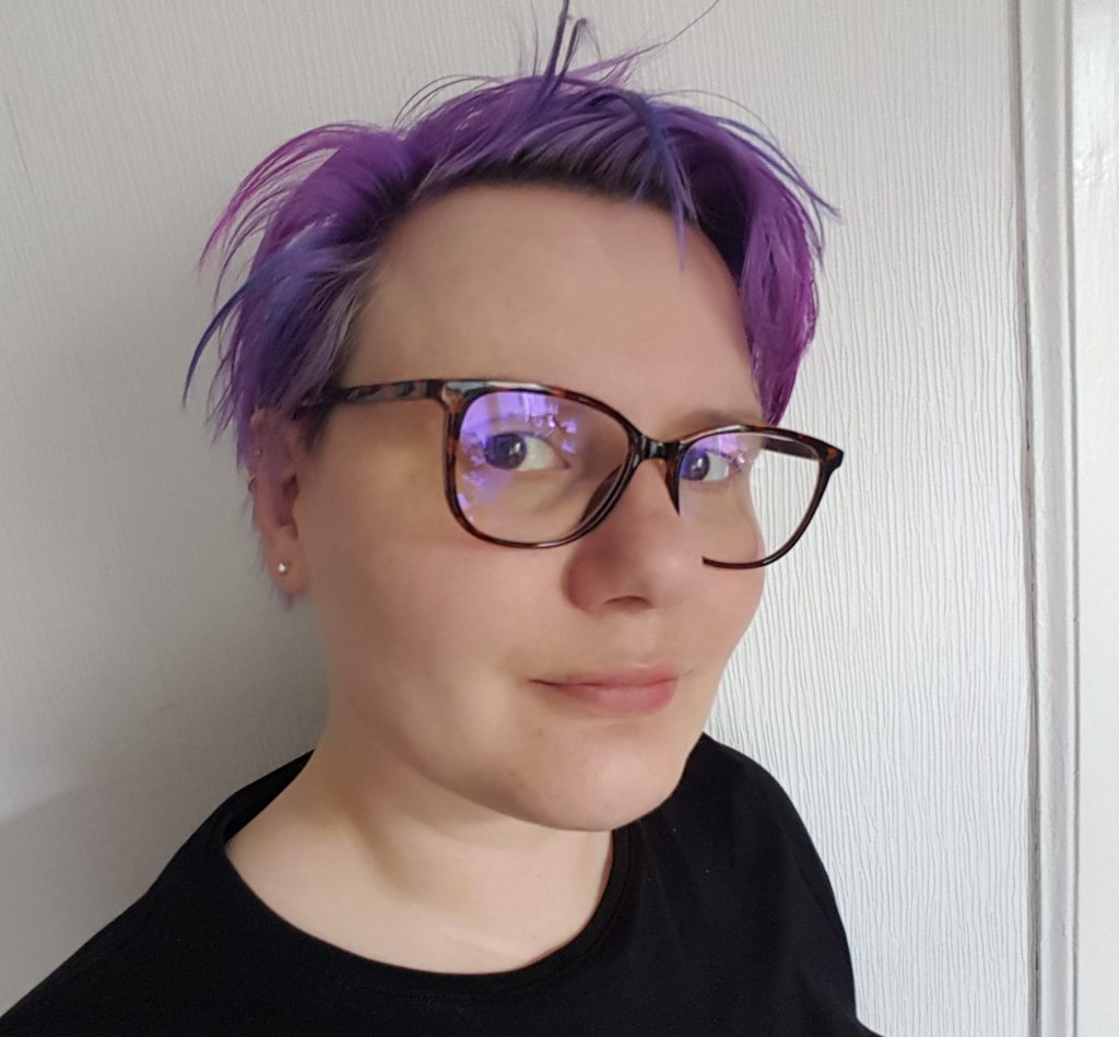 A brunette white woman with purple hair a tortoiseshell glasses wearing a black top.
